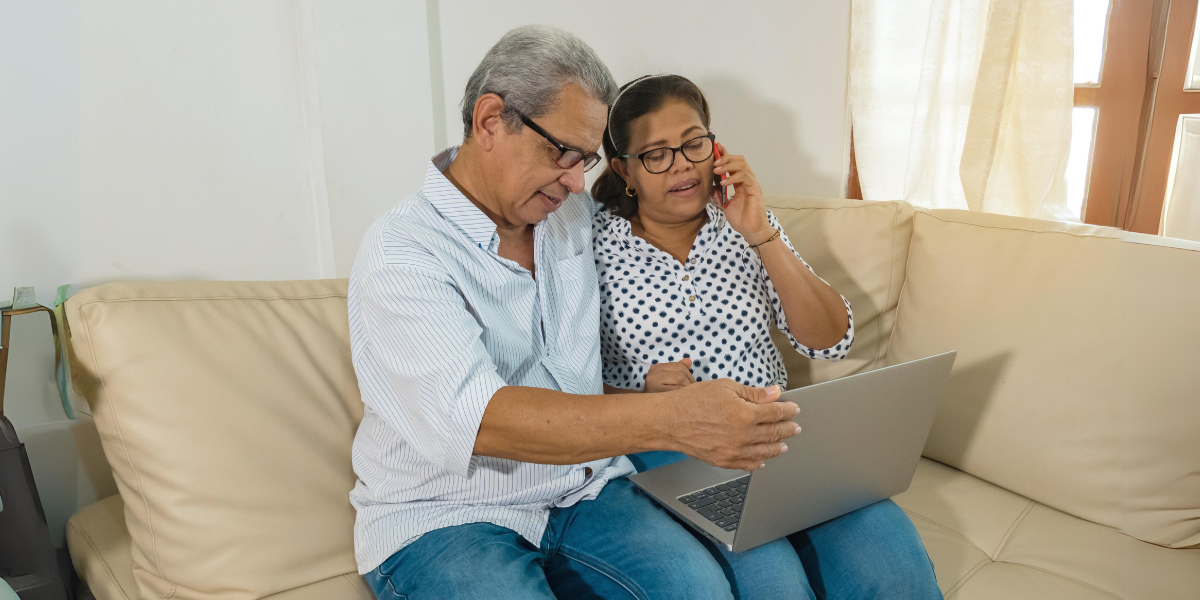 Older Latino couple sits on sofa looking at computer